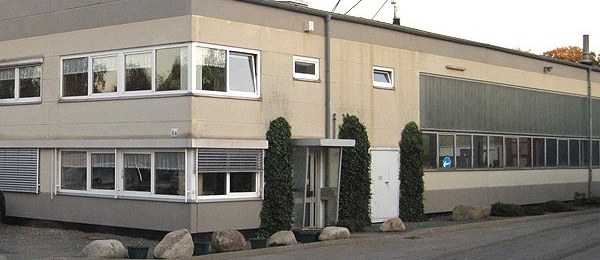 Zweigeschossiges Bürogebäude mit umlaufendem Fensterband, daran anschließend die Hallenwand; vor dem Eingang schmale Säulenbäume, am Straßenrand große Findlinge.